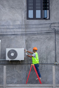 Worker in hard hat and high visibility vest installs or repairs outdoor air conditioning unit on gray building wall, standing on red ladder.