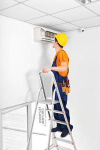 Man wearing a hard hat, overalls, and toolbelt standing on a ladder to inspect a wall-mounted air conditioning unit