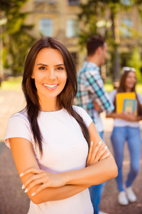 Smiling young woman with crossed arms standing in front of a university campus with students in background