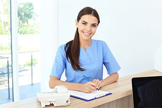 Smiling female medical student in blue uniform writing on a clipboard at a front reception desk.