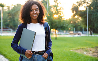 Young woman with curly hair holding a laptop, standing in a park at sunset.