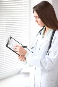 Doctor in white lab coat with stethoscope reading a document on a clipboard by a window with blinds.