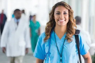 Nurse in blue scrubs with a stethoscope and backpack standing in a hospital hallway with blurred healthcare professionals in the background.