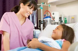 Nurse comforting young girl in hospital bed, both smiling, in a medical setting with equipment in the background.