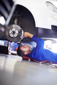 Mechanic in uniform working underneath car on a creeper, repairing or inspecting vehicle's suspension and brakes at auto service garage.