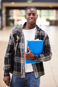 Young male student smiling, holding blue folders and wearing a plaid shirt with a backpack in a school corridor.