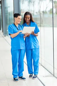 Two nurses in blue scrubs discussing over a digital tablet in a hospital corridor