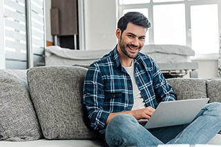 Happy young man using a laptop while sitting on a gray couch in a living room.