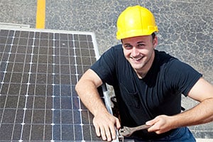 Tradesperson in a yellow hard hat and black shirt, smiling while adjusting a solar panel with a wrench.