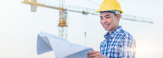 Smiling young man in a plaid shirt and yellow hard hat looking at paper blueprints in front of a construction crane outside