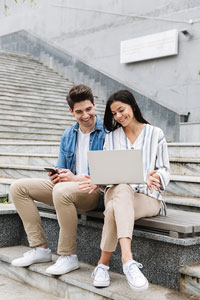 Smiling young man and woman sitting on concrete steps outside and looking at an open laptop together